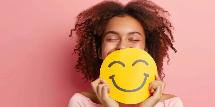 woman holding up an emoji face on pink background, smiley happy person smiling and showing emoticon symbol with eyes closed