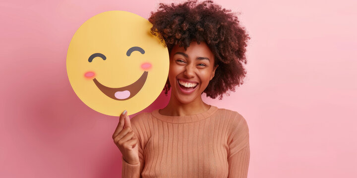 woman holding up an emoji face on pink background, smiley happy person smiling and showing emoticon symbol with eyes closed