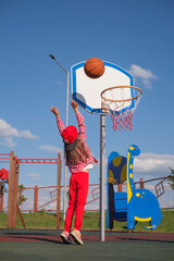 A little girl in a cap and a Checkered shirt throws a ball into a basketball hoop on the playground.
