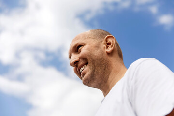 Bald man in white t-shirt smiling, looking down, blue sky