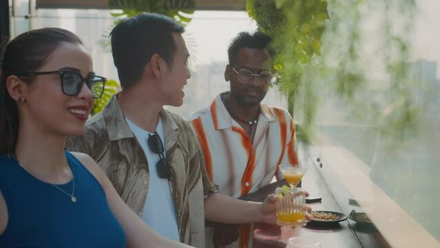 Side view of diverse coworkers spending time in fancy bar with cocktails and talking near panoramic window