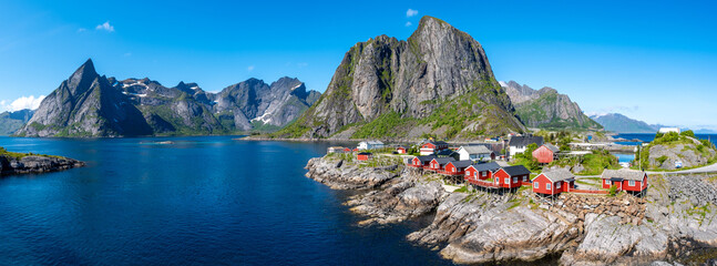 A picturesque view of red cabins perched on the rocky shore of a fjord in Norway, with towering...