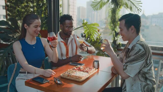 Group of bicultural friends toasting with cocktails while sitting at table with pizza at rooftop cafe