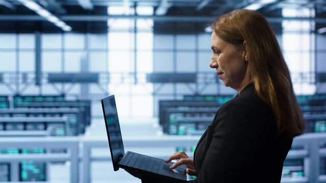 System administrator in server hub checking recovery plan on laptop, monitoring energy consumption. IT expert uses notebook in data center workspace, ensuring rigs are functioning optimally, camera A