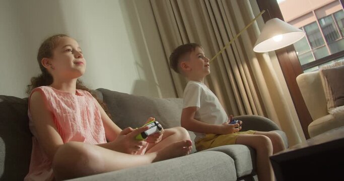 Cute little girl and boy smiles, kids are playing video games on the tv, actively plays computer games sitting on sofa in living room. Children playing computer games. Children enjoy multimedia games.