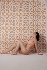 A beautiful slender girl in pink swimsuit in a Moroccan-style sand studio. A pretty brunette lady...