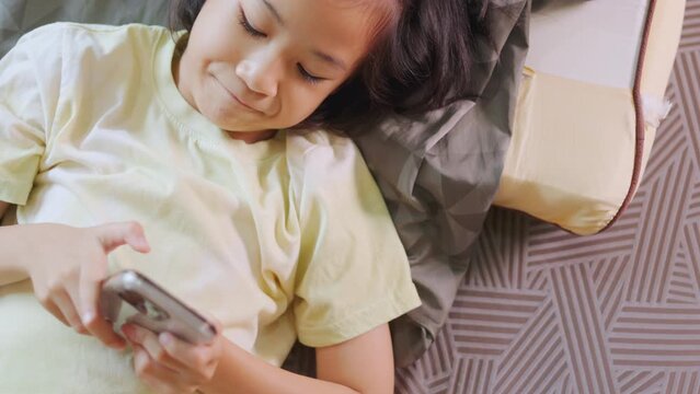 Top view of adorable Asian kid girl, lied in bed, enjoys her smartphone in a sunlit room. Her expression hints at digital fun, highlighting the allure of home tech, playing game and e-learning.