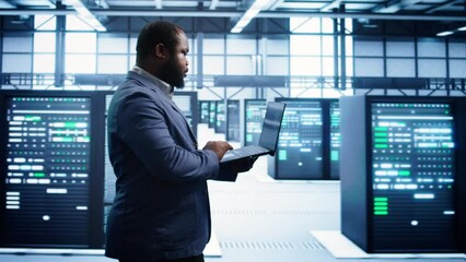 African american man walking in high tech facility, using laptop to patch network issues leading to slow data transfer rates. Data center worker fixing errors affecting supercomputers, camera B