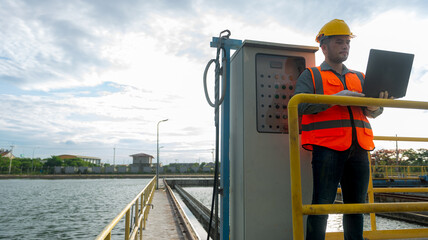 an engineer stand in front of  Pump control cabinet while holding laptop on his left hand and checking value of the gauge at waste water treatment