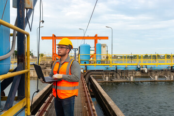 an engineer stand beside the blue water tank while his right  hand holding laptop his left hand...