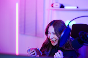 Asian excited woman streaming in LED neon light room holding a phone while checking championship arena win score result surrounded by gaming and tech equipment