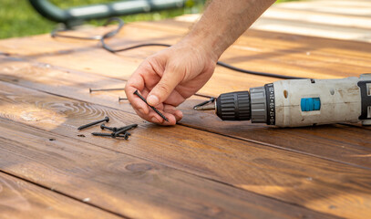 A close-up shot of a hand holding a screw and a cordless drill lying on a wooden table. Scattered...