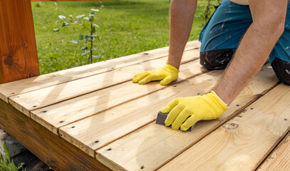 A close-up shot of a persons hands wearing yellow gloves, sanding a wooden deck with sandpaper on a sunny day.