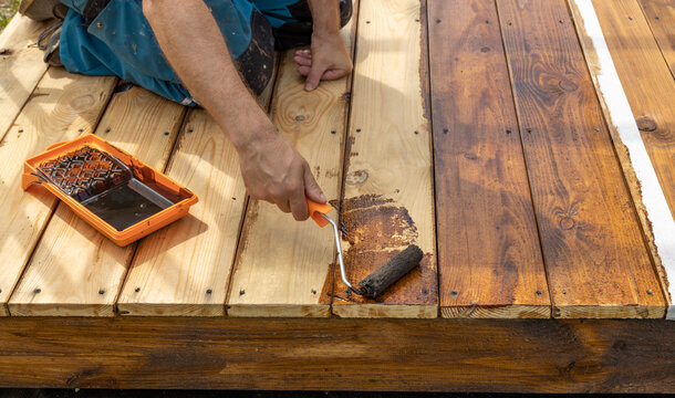 A person is staining a wooden deck with a brown stain using a paint roller. The deck is partially stained with a visible difference in color.