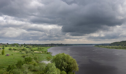A view of a wide, calm river flowing through a verdant landscape, with a dramatic sky filled with...