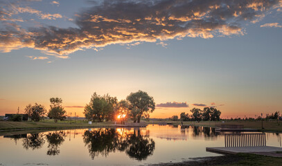 A serene sunset paints the sky with hues of orange and pink, reflecting off the tranquil waters of a park lake. Trees line the shore, creating a picturesque backdrop for a peaceful evening.
