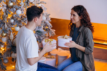 Young couple is seen exchanging gifts while sitting by beautifully decorated Christmas tree with golden and white ornaments. Scene is cozy and festive, with warm lighting and fireplace in background. 