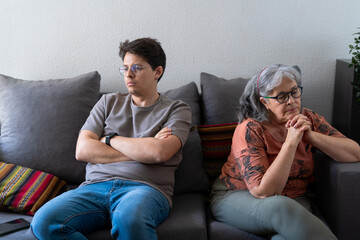 A mother and her adult son sit on a couch, visibly upset after an argument. The son has his arms...