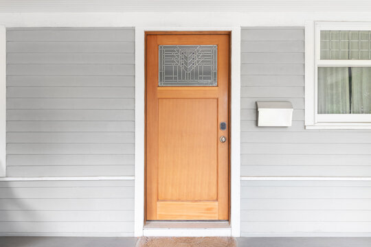 A wooden front door with a glass window on a grey and white homes covered front porch.
