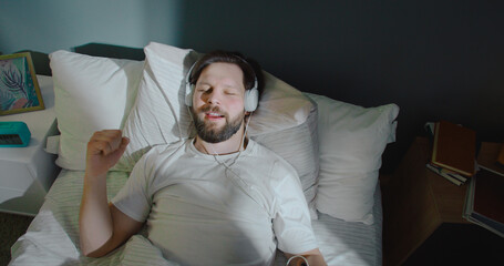 Dreamy attractive young man lying in bed, smiling and listening to music in headphones. Feeling...