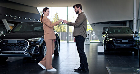 Confident young caucasian man and dealer in Covid-19 face masks shaking hands and passing keys. Young businessman is buying automobile in auto dealership on pandemic.