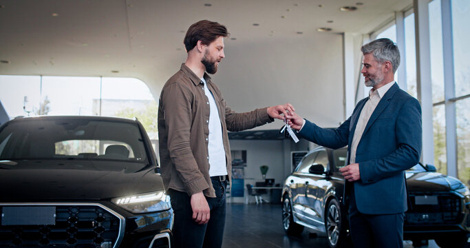 Caucasian car consultant presentating keys from new car to male client at salon. Happy young guy buying new transport. Selling and business concept.