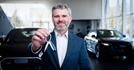 Portrait of smiling car dealer in formal suit and glasses holding keys from new car. Male dealer standing at auto salon, smiling and looking at camera. Selling and business concept.