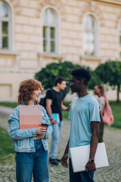 Diverse exchange student with laptop chatting with classmate at campus