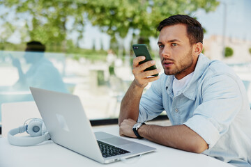 Man in casual clothing using a smartphone for voice command while working on a laptop outdoors.