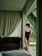 Obraz premium Young woman in black swimsuit standing by large window, looking outside with thoughtful expression in modern living room
