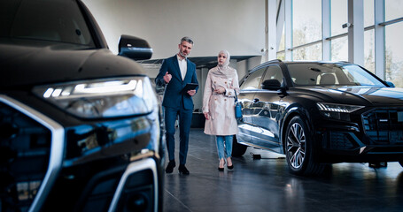 Professional car salesman is telling interested buyer about luxurious car in motor show while Arabian woman is looking at auto and listening to dealer. Manager in auto dealership presents new car.