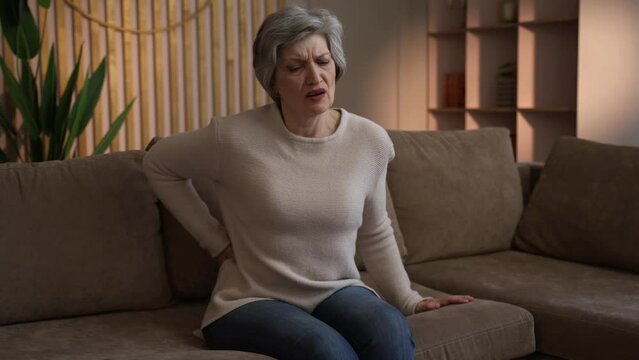 An elderly woman, getting up from the sofa, experiences back pain. She sits on the couch and reaches for a pillow to help herself up, but her back starts to hurt. She sits back down with a grimace, as