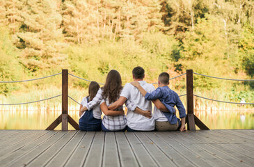 Beautiful family sitting on a pier with view lake. A family hugs outdoor while looking at the water