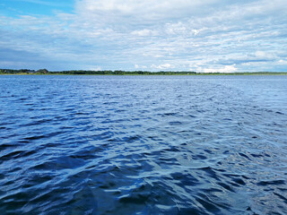 A calm lake with a blue water surface with waves, against the background of a green forest with trees and plants on the horizon under clouds and a blue sky. Environment, natural landscape, travel