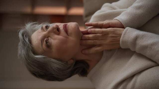 Vertical Screen: An elderly woman checks her thyroid gland at home. She is sitting on the couch and looking in front of her. Her hands touch the front of her neck, next to the thyroid gland
