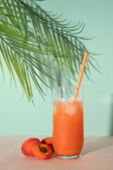 Healthy refreshing summer cocktail drink with apricot fruit juice, ice and drinking straw on pastel peachy pink and turquoise blue background with soft sunlight shadow and palm tree leaves