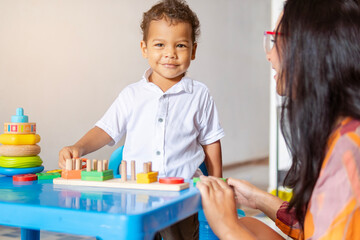 Niño latino de 2 años en compañía de su madre mientras juega en una mesa azul con diferentes juegos prácticos y didácticos. Están en su casa, ambos son latinos