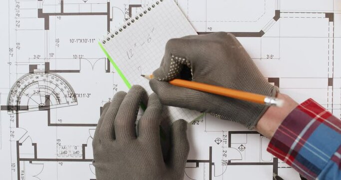 Engineer writes marks in notepad measuring floor plan with protractor view from above. Designer checkup drawing of building in office