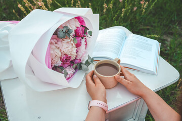 A bouquet of peonies, an open book and a cup of tea in hands, a pleasant time in nature