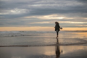 Naklejka premium Woman walking along the beach at sunset, holding her shoes in her hand