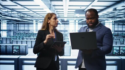 Teamworking colleagues in server farm using laptop to patch network issues affecting transfer rates. Coworkers in data center doing brainstorming, fixing errors affecting supercomputers, camera B