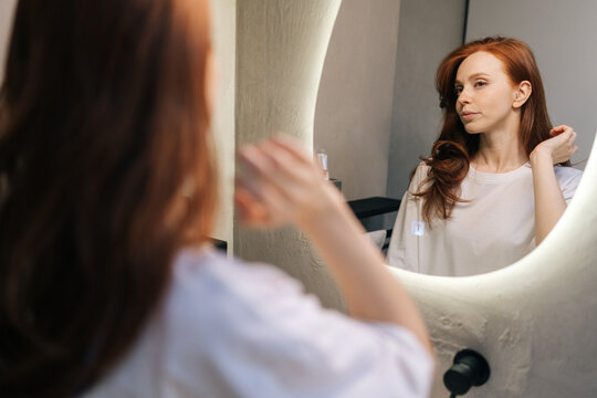 Beautiful young woman in casual clothes, preening, do morning routine, touch her hair standing in front of mirror, smiling admire herself feels satisfied with appearance and complexion.