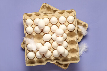 Cardboard box with white chicken eggs and feathers on lilac background