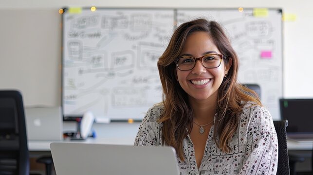 Smiling professional woman working on a laptop, whiteboard in background, office environment