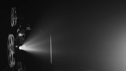 A black-and-white vintage movie projector working in the dark. A bright beam of light shines through the lens, illuminating the film reels. The atmosphere evokes classic film screenings