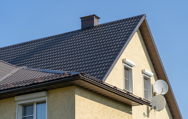 Close-up shot of a house with a black metal roof, a chimney, and two windows on the side. The house is painted a pale yellow