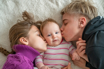 Baby girl lies in middle of a bed, surrounded by two older siblings. The older siblings are kissing babys cheeks