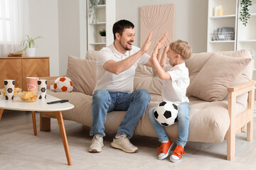 Young father and little son with soccer balls sitting on sofa at home