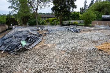 New home foundation construction in progress, gravel work road onto job site, cement retaining walls in place, black tarp covering dirt backfilled into foundation form, residential building site
