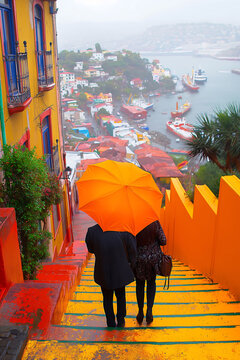 A couple with an umbrella walking down the stairs in a Valparaiso-like city with colorfully painted houses on a cloudy day, creating a vibrant yet atmospheric scene